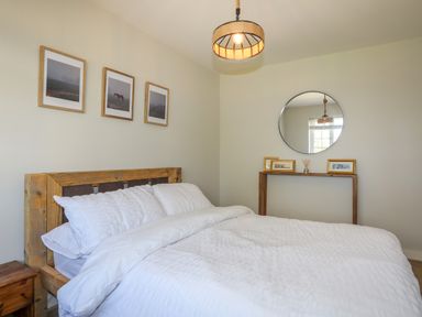 A bedroom with a wooden bed frame white bedding three pictures above the bed a round mirror on the wall and a wooden console table with framed pictures at Iscoed in Llangefni