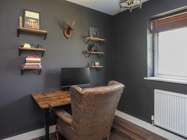 A home office with a wooden desk a computer monitor a brown upholstered chair shelves with books and decorations and a window with blinds at Iscoed Llangefni