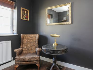 A corner room with a patterned armchair a round wooden table with a brass lamp a large framed mirror and a small framed painting at Iscoed in Llangefni