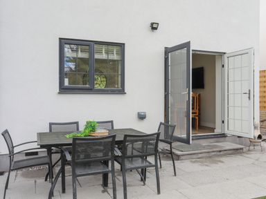 An outdoor patio with a black metal table and six chairs next to a gray wall with a window and an open door at Iscoed in Llangefni