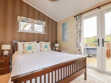 A bedroom with a wooden bed frame two bedside tables with lamps and patio doors leading outside at Hideaway Lodge Bossiney near Tintagel
