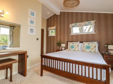 A bedroom with a wooden bed two floral pillows two bedside tables with lamps a dressing table with a stool and three framed pictures on the wall at Hideaway Lodge in Bossiney near Tintagel