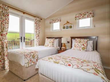 A bedroom with two single beds a nightstand and lamp with floral curtains and cushions at Hideaway Lodge in Bossiney near Tintagel