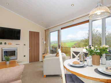 A living room with dining table set with cups and bread and a large window showing green hills at Hideaway Lodge Bossiney near Tintagel