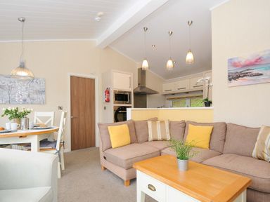 A living room with a brown sectional sofa and yellow cushions a wooden coffee table and a dining table with chairs at Hideaway Lodge in Bossiney near Tintagel