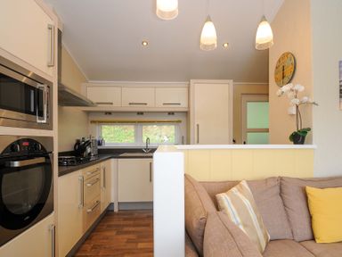 A kitchen with built-in oven and microwave a stove refrigerator and a sink next to a living area with a sofa and cushions at Hideaway Lodge in Bossiney near Tintagel
