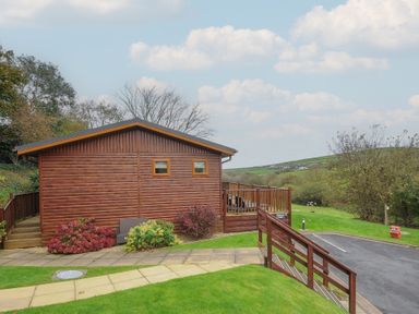 A wooden cabin with a deck and garden path surrounded by grass and shrubs at Hideaway Lodge Bossiney near Tintagel