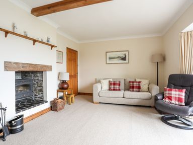 A living room with a beige sofa and red plaid cushions next to a black leather armchair with a red plaid cushion a fireplace with a wooden mantel and a basket of firewood beside a wooden door and a lamp on a small table at Bwthyn in Borth-y-Gest near Porthmadog