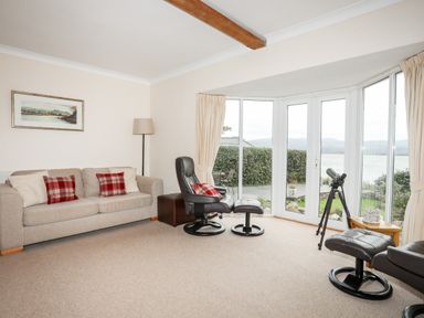 A living room with a beige sofa and leather chairs near large windows overlooking water at Bwthyn in Borth-y-Gest near Porthmadog