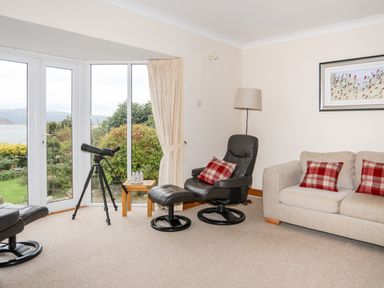 A living room with a telescope near bay windows a black leather chair with an ottoman and a beige sofa with red plaid cushions at Bwthyn in Borth-y-Gest near Porthmadog
