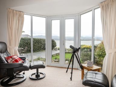 A sitting area with two black leather chairs a wooden side table a telescope and large windows overlooking a garden and water at Bwthyn in Borth-y-Gest near Porthmadog