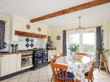 A kitchen with a round dining table and wooden chairs next to a stove and oven with patterned curtains at Bwthyn in Borth-y-Gest near Porthmadog