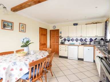 A kitchen with a dining table and wooden chairs a dishwasher a fridge and white cabinets at Bwthyn in Borth-y-Gest near Porthmadog