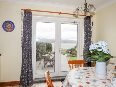 A dining area with table and chairs facing glass doors leading to a patio with table and chairs and a view of water and hills at Bwthyn in Borth-y-Gest near Porthmadog