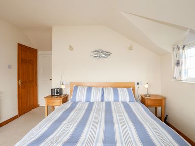 A bedroom with a double bed with blue and white striped bedding two wooden bedside tables and a window with patterned curtains at Bwthyn in Borth-y-Gest near Porthmadog