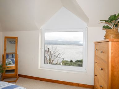 A bedroom with a wooden dresser a standing mirror and a window showing a lake and hills at Bwthyn Borth-y-Gest near Porthmadog