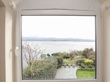 A window view showing a garden with shrubs and potted plants beside a patio and a water body with distant hills at Bwthyn in Borth-y-Gest near Porthmadog