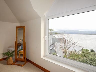 A corner of a room with a standing mirror, a plant, and a large window overlooking water and rooftops at Bwthyn in Borth-y-Gest near Porthmadog