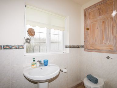 A bathroom with a white sink holding a soap dispenser and blue cup a wooden cabinet above a toilet with a folded towel and a window with a roller blind at Bwthyn in Borth-y-Gest near Porthmadog