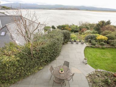 A patio with a round table and chairs near a garden with bushes and grass overlooking a body of water at Bwthyn in Borth-y-Gest near Porthmadog