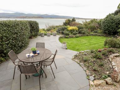 An outdoor patio with a round table and four chairs overlooking a garden and water at Bwthyn in Borth-y-Gest near Porthmadog