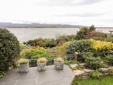 A patio with three square planters overlooking a garden with shrubs and a body of water beyond at Bwthyn in Borth-y-Gest near Porthmadog