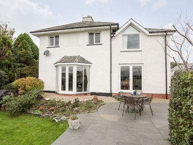 The back of a two-story white house with a garden patio with a round table and four chairs and some plants and bushes at Bwthyn in Borth-y-Gest near Porthmadog
