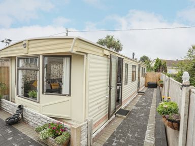 An exterior view of a beige mobile home with a paved walkway and flower pots along the fence at Rhiwlas Bach in Benllech