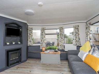 A living room with a gray sofa yellow and white pillows a small table with a lamp books and plants a television above a fireplace and patterned curtains at Rhiwlas Bach in Benllech