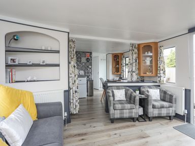 A living room with two plaid armchairs a gray sofa with yellow and white pillows shelves with pictures a small kitchen area with patterned wall tiles and wooden cabinets at Rhiwlas Bach in Benllech