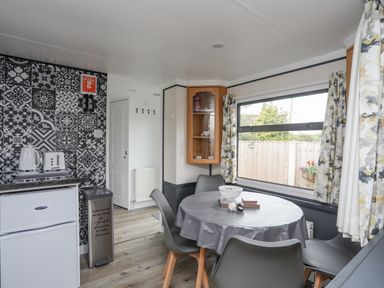 A dining area with a round table and four chairs next to a large window and a small kitchen corner with a fridge toaster kettle and a patterned tiled wall at Rhiwlas Bach in Benllech
