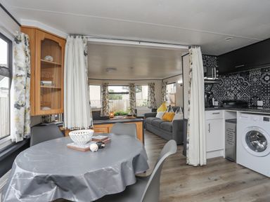 A dining area with a round table and chairs next to a kitchen with washing machine and black patterned backsplash at Rhiwlas Bach in Benllech