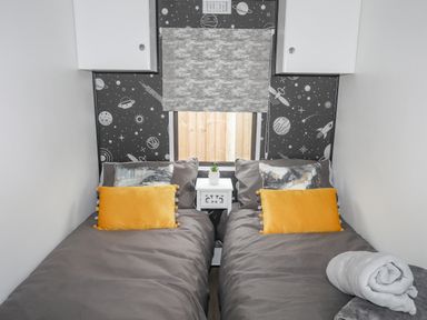 A bedroom with two single beds gray bedding yellow pillows a nightstand and space themed wallpaper at Rhiwlas Bach in Benllech