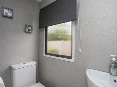 A bathroom with a toilet a small sink a window with a dark roller blind and framed wall decorations at Rhiwlas Bach in Benllech