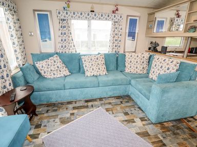 A living room with a blue corner sofa floral curtains and cushions wooden side table and patterned rug at Beachcomber D35 in Towyn