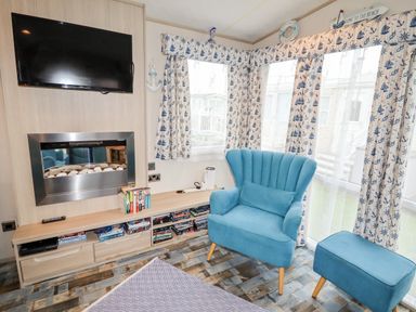 A sitting area with a blue armchair and ottoman near windows with nautical curtains a wall-mounted television and fireplace at Beachcomber D35 in Towyn