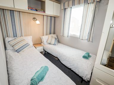 A bedroom with two single beds a bedside table a window with curtains and a wardrobe at Beachcomber D35 in Towyn