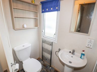 A small bathroom with a toilet, sink with soap, mirror, window with a blue blind, wall shelves, and a towel radiator at Beachcomber D35 in Towyn
