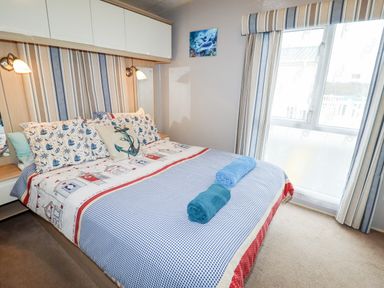 A bedroom with a double bed with nautical themed bedding two rolled towels on the bed striped curtains and a window at Beachcomber D35 in Towyn