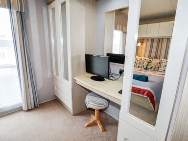 A bedroom with a desk, TV, stool, wardrobe, and a bed reflected in a mirror at Beachcomber D35 in Towyn