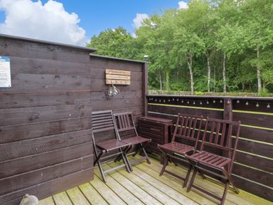 An outdoor wooden deck with four wooden chairs a small wooden storage box and a sign on the wall surrounded by trees at Glenisle in Dalbeattie