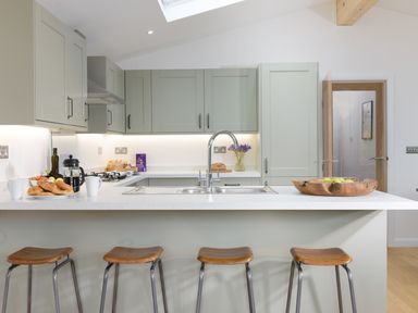 A kitchen with a countertop island with four stools a sink and faucet croissants and mugs on the counter and cabinets with under cabinet lighting at 4 Valley View in Lanreath