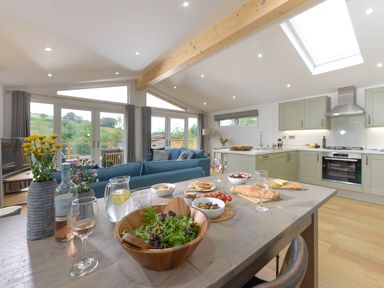 A kitchen and dining area with a wooden table set with food and drinks a blue sofa and large windows in the background at 4 Valley View in Lanreath