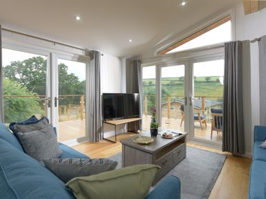 A living room with two blue sofas a wooden coffee table a TV on a stand and large windows overlooking a deck and greenery at 4 Valley View in Lanreath