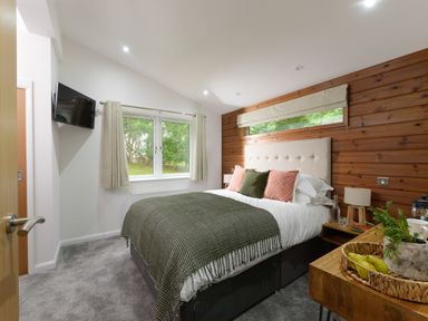 A bedroom with a bed with pillows and blankets a window with curtains a wooden wall and a wooden table with a plant and fruit basket at 4 Valley View in Lanreath