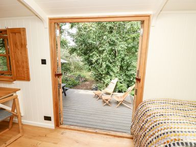 A bedroom with a bed covered in a patterned blanket opening onto a wooden deck with two wooden lounge chairs and green trees outside at The Happy Hut in Sopley near Christchurch