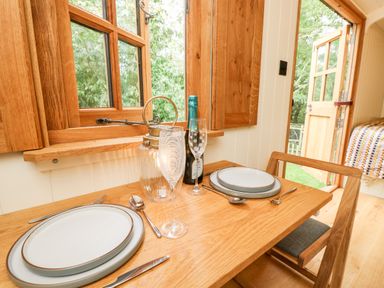 A wooden dining table with two place settings near an open window and door with a bottle and glasses at The Happy Hut in Sopley near Christchurch
