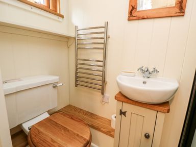 A bathroom with a toilet with wooden seat a small white sink on a wooden cabinet a wall-mounted chrome towel rack and a wooden framed mirror at The Happy Hut in Sopley near Christchurch