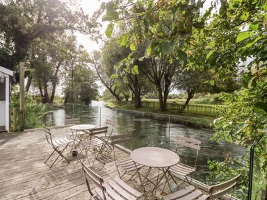 A wooden deck with metal tables and chairs overlooking a river and trees at The Happy Hut in Sopley near Christchurch