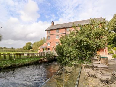 An outdoor seating area with tables and chairs next to a river and a brick building with a slate roof at The Happy Hut in Sopley near Christchurch